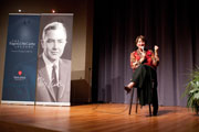 A person sits on a chair on stage, speaking passionately into a microphone. The backdrop is a large curtain with a banner featuring a black-and-white portrait of an older man in a suit. There's a small plant on the right side of the stage.