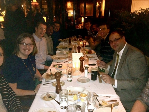 A group of people sitting around a long dining table in a dimly lit restaurant. They are smiling and enjoying a meal together, with plates and drinks on the table. The setting is cozy and social.