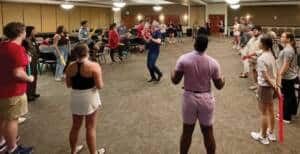 A group of people stands in a circle in a conference room, participating in an interactive activity. Some hold colorful sticks. Chairs are arranged along the walls, and the atmosphere appears lively and engaging.