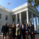 A group of people stands in front of the White House. They are posing for a photo on a sunny day, with the American flag visible on top of the building. The group consists of both men and women dressed in formal attire.