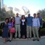 A group of eight people stand closely together, smiling in front of the metallic structure of the Pritzker Pavilion in Millennium Park, Chicago. The sky is clear and trees are in bloom around the area.