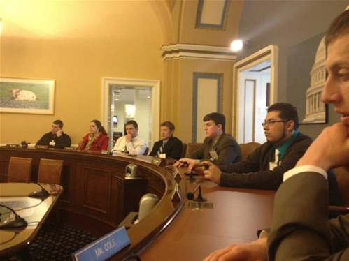 A group of people seated around a large wooden conference table in a formal room. The room features ornate decor and a painting on the wall. A name plaque reading "MR. COLE" is visible on the table in the foreground.