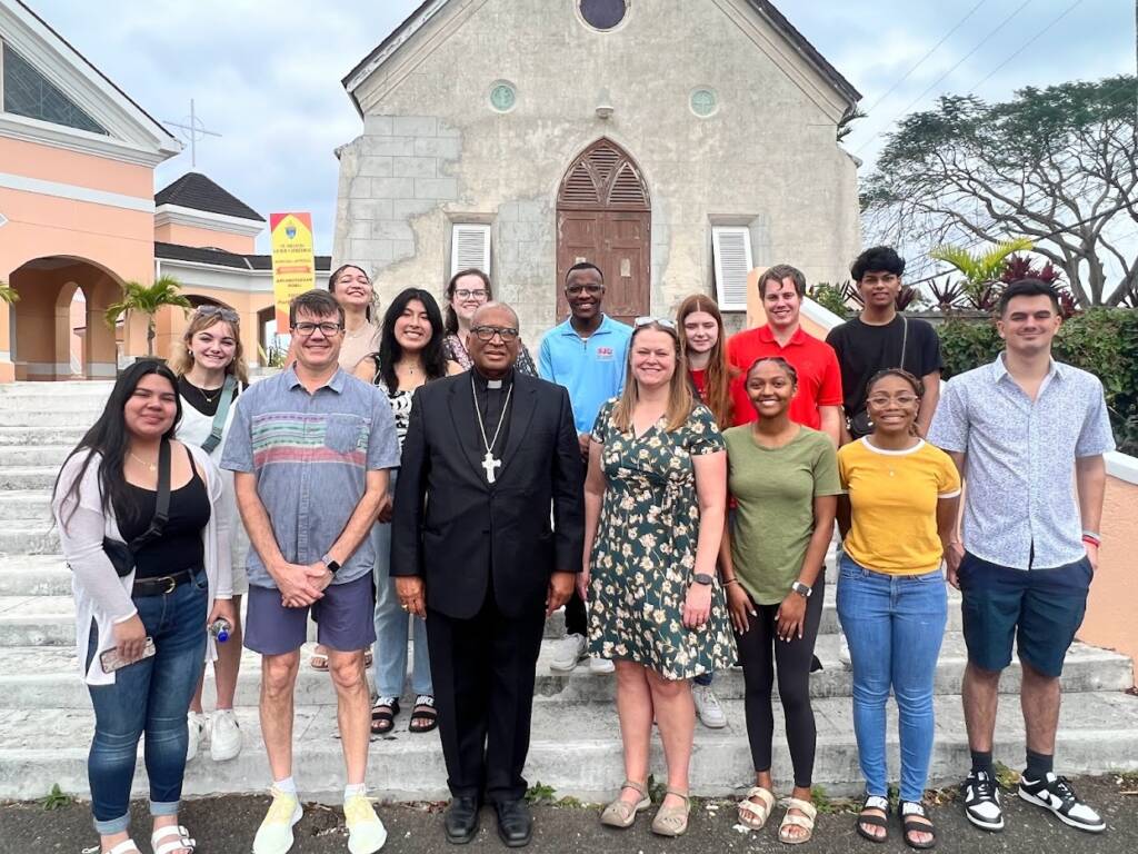 A group of people, including a person in religious attire, stand on the steps of a church. They are all smiling and posing for the photo in front of a stone building with arched windows. The sky is overcast, and there are trees in the background.