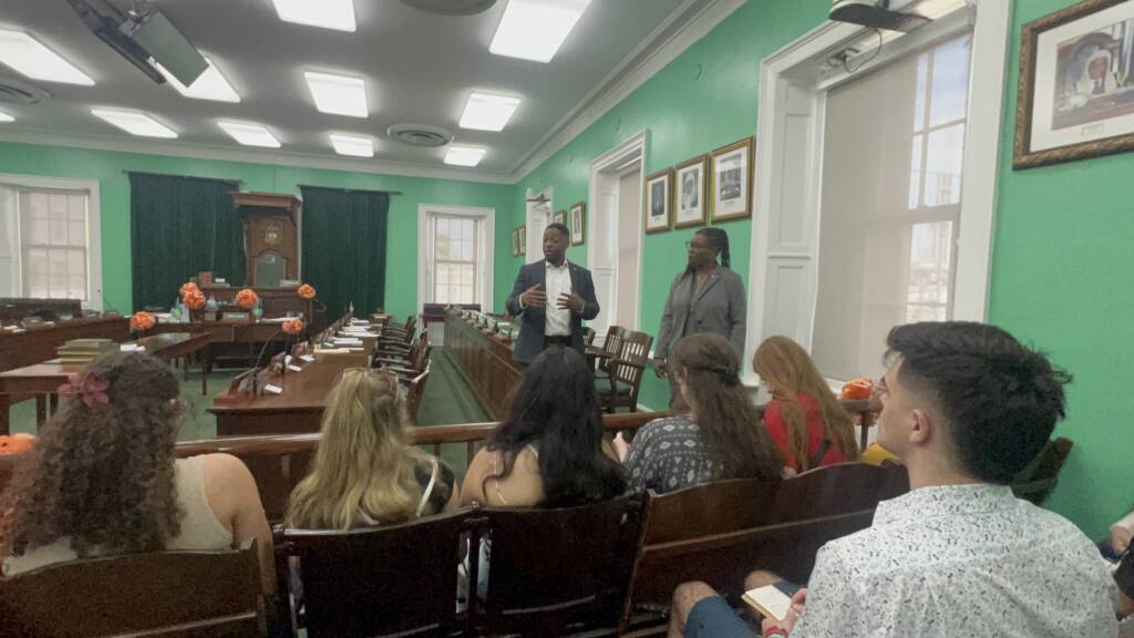 A group of people sit in a room with green walls, listening to a speaker standing at the front. Another person stands nearby. The room has several portraits and decorations, and windows let in natural light.