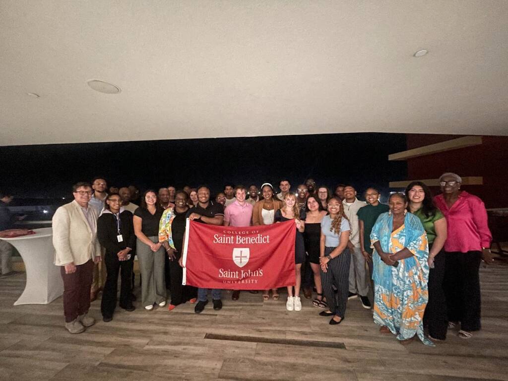 A large group of people gather indoors, smiling and holding a red banner that reads "College of Saint Benedict" and "Saint John's University." The setting appears to be a room with wooden floors and dim lighting.