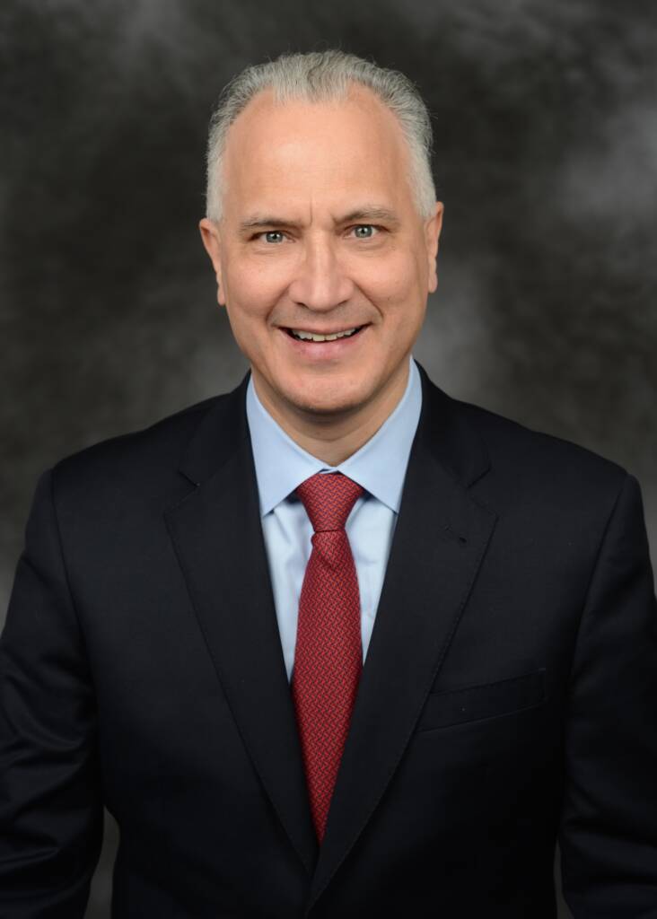 A man with short gray hair is wearing a dark suit, light blue shirt, and red tie, smiling against a gray background.