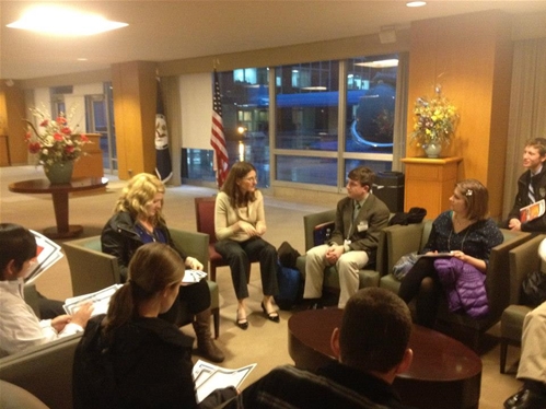 A group of people sitting in a circle in a waiting room, engaging in a conversation. The room has a modern, professional feel with potted plants and flags in the background. Some people hold papers as they listen attentively.