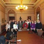 A group of people posing for a photo in an ornate room with decorative ceilings and a chandelier. They are standing behind a large table with several chairs around it. A bust is visible in the background.