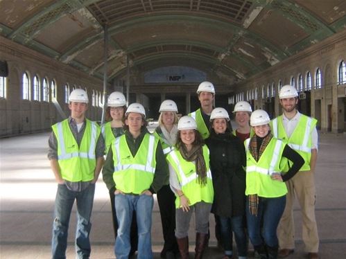 A group of ten people wearing yellow safety vests and white hard hats pose together inside a large, empty building with high ceilings and arched windows.