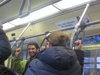 Passengers standing and holding onto handrails inside a bus or tram. A man in a green jacket is smiling. An advertisement on the wall reads, "Pull the trigger on savings." The interior appears crowded.