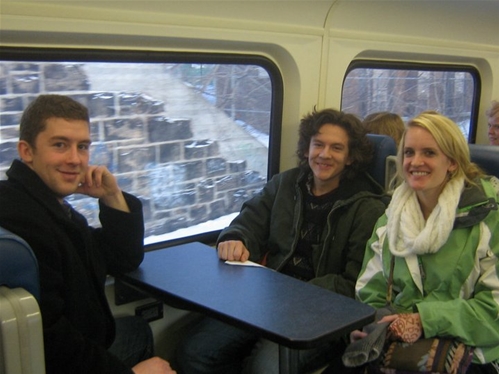 Three people sit at a table on a train, with a man and woman smiling and another man looking at the camera. They are dressed in casual winter clothing. The train window shows a blurred outdoor landscape with trees and a stone wall.