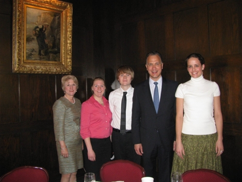 A group of five people posing together in a room with dark wood-paneled walls. They are dressed in semi-formal attire. A framed painting hangs on the wall above them.