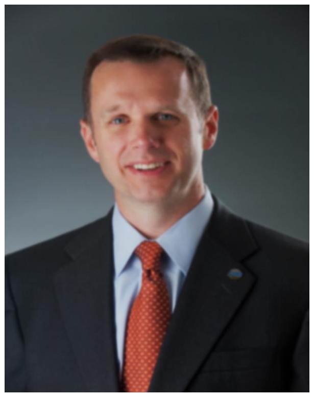 A professional portrait of a man wearing a dark suit, light blue shirt, and orange tie, smiling slightly against a plain dark background.