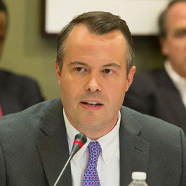A man in a suit and tie speaking into a microphone at a conference or meeting. Other people are blurred in the background.