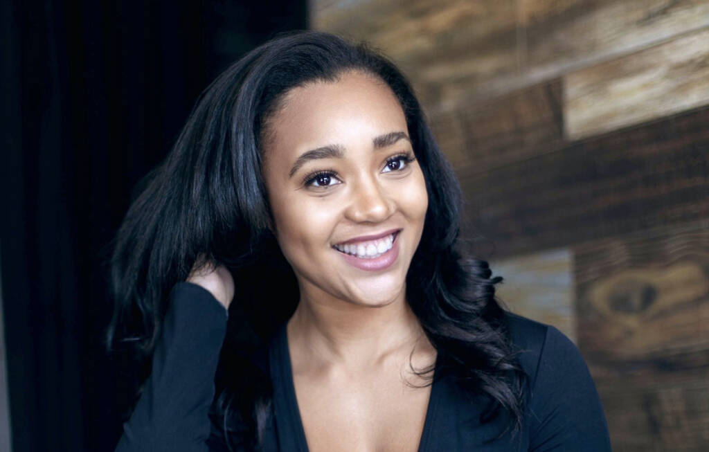Smiling woman with long, dark hair touches her hair in a relaxed pose. She wears a black top, and the background features wooden paneling.