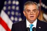 A man in a suit and blue tie speaks at a podium. American flags are visible in the blurred background.