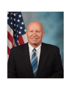 U.S. Rep. Kevin Brady. A bald man in a suit and blue striped tie poses for a professional portrait against a backdrop of the American flag and a blue and gray gradient background.
