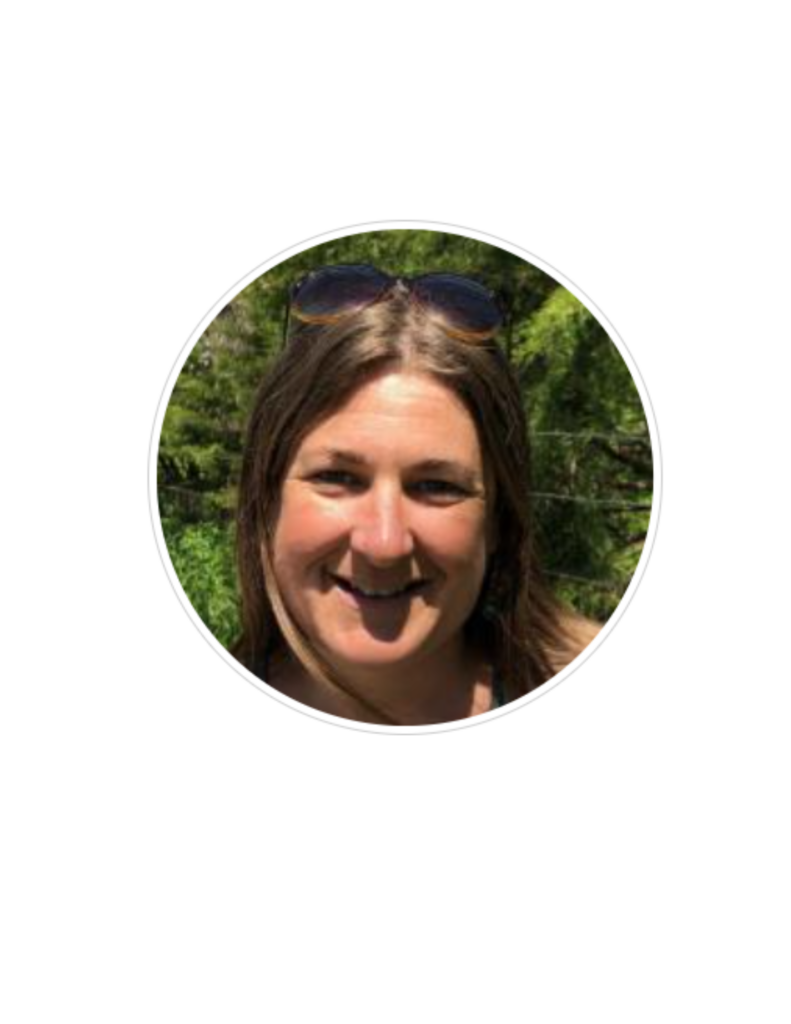 A person with long brown hair is smiling outdoors. They are wearing sunglasses on their head. The background shows greenery and trees.