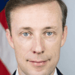 A man with light skin, light brown hair, and blue eyes wearing a suit and tie stands in front of a blurred American flag, prepared to speak at the 19th Annual McCarthy Lecture.