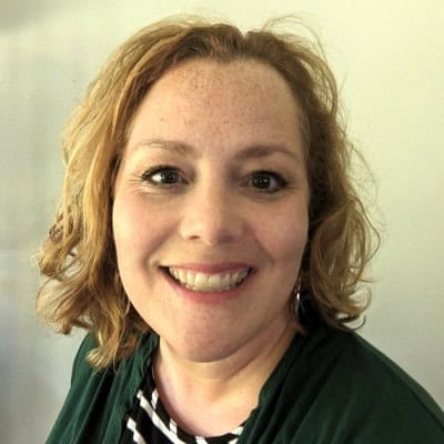 A woman with curly, shoulder-length blonde hair smiles at the camera. She is wearing a green top over a black and white striped shirt, with a neutral background behind her.