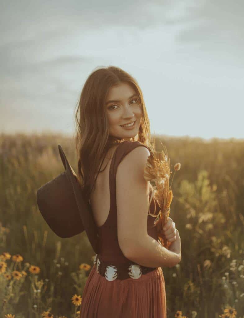 A woman stands in a sunny field of wildflowers, holding dried grasses. She wears a brown dress, a wide belt, and a black hat hanging down her back, smiling and looking over her shoulder at the camera.