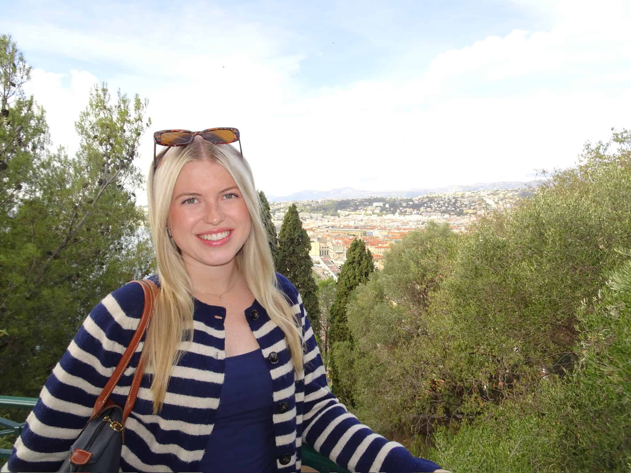 A smiling woman with long blonde hair, wearing a navy shirt and striped cardigan, stands outdoors with sunglasses on her head. Behind her are trees and a scenic cityscape under a partly cloudy sky.
