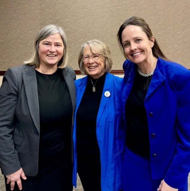 Three women standing together and smiling. Two women are wearing blue blazers and one is wearing a gray blazer. They are indoors in front of a beige wall.