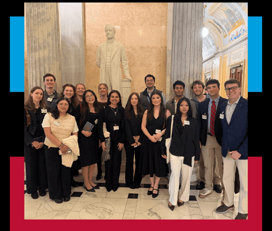 A group of fifteen people dressed in business attire pose for a photo in front of a white statue inside a grand building with marble columns and ornate ceilings.