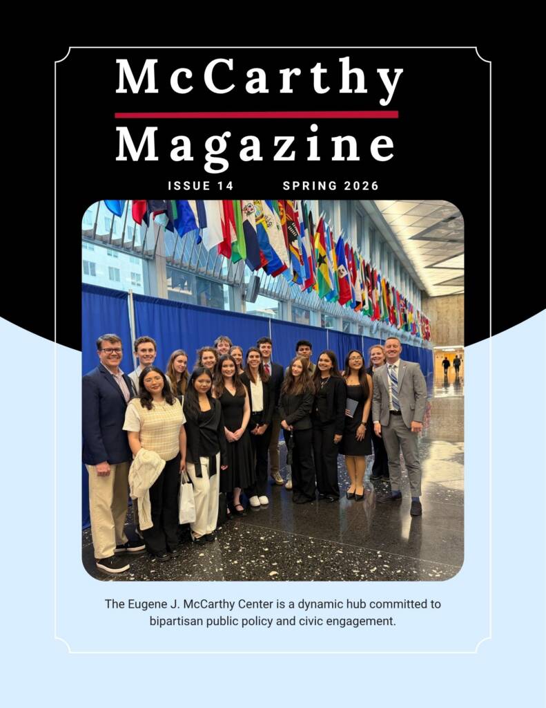 A group of professionally dressed people poses and smiles together inside a modern building adorned with rows of colorful international flags overhead. The text identifies the image as the cover of McCarthy Magazine, Spring 2026, Issue 14.