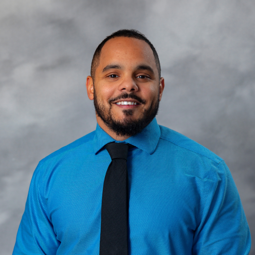 A man with short hair and a beard is smiling while wearing a bright blue shirt and a black tie. The background is a neutral gray with a soft focus.
