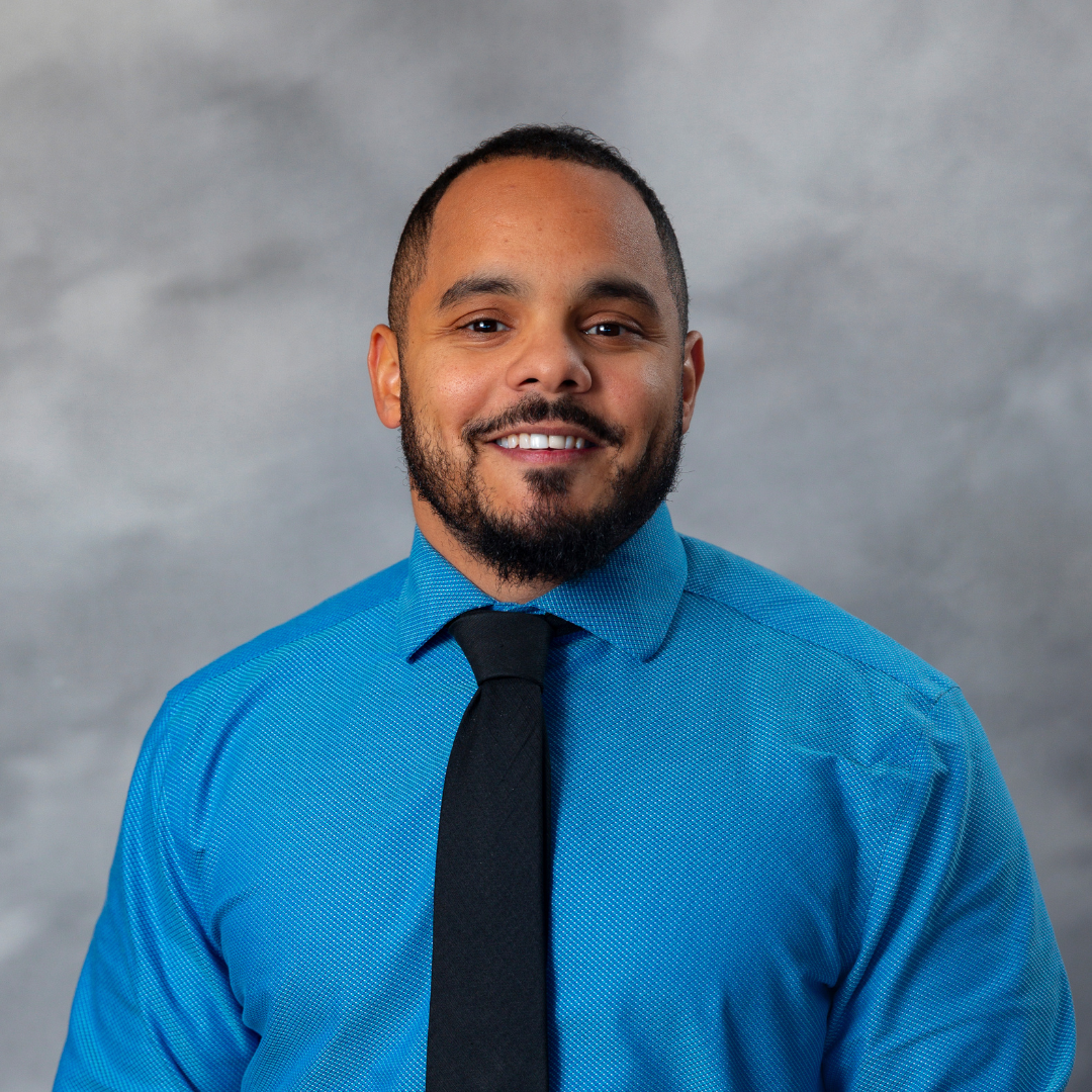 A man with short hair and a beard is smiling while wearing a bright blue shirt and a black tie. The background is a neutral gray with a soft focus.