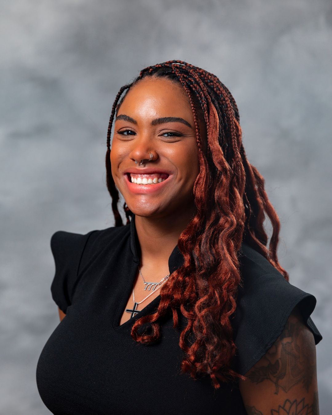 A person with braided hair smiles warmly, wearing a black top. The background is a soft gray.