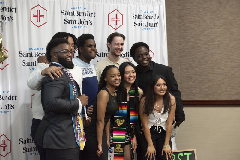 A group of nine young adults, dressed in semi-formal and festive attire, smile and pose together in front of a College of Saint Benedict and Saint John’s University backdrop.