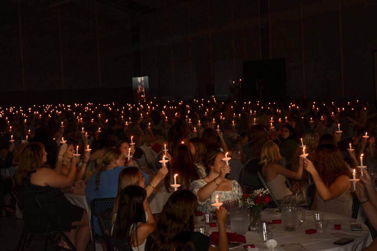 A large group of people seated at tables in a dimly lit room, each holding a lit candle, creating a warm glow throughout the space.