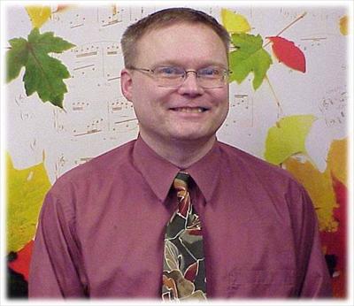 A man with short hair and glasses is smiling. He is wearing a maroon shirt and a tie with a leaf pattern. The background features autumn leaves and music notes.