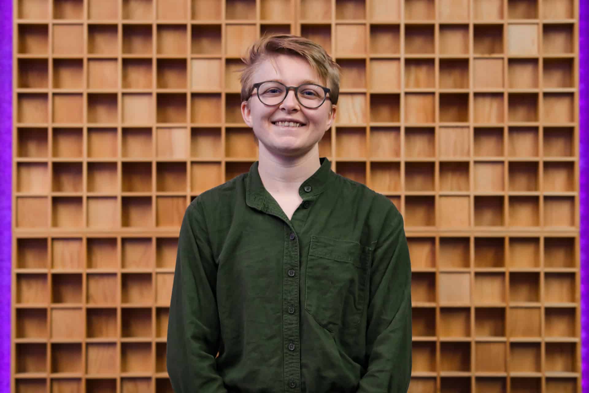 A person with short, light hair and glasses, wearing a green button-up shirt, stands smiling in front of a wooden grid-patterned wall.