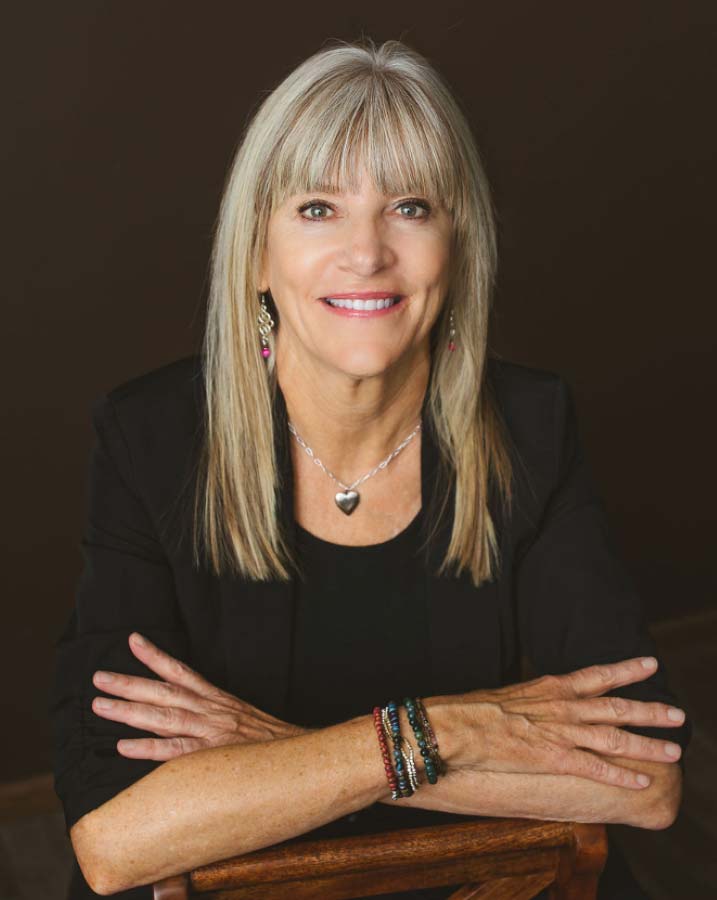 A woman with long, straight gray hair and bangs smiles warmly. She is wearing a black top and a black blazer, accessorized with a silver heart necklace and dangly earrings. Her arms are crossed and she has multiple bracelets on her right wrist. The background is dark.