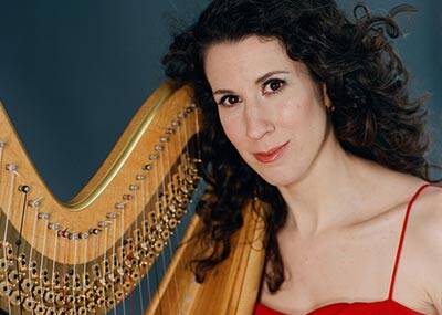 A woman with curly dark hair poses next to a harp, wearing a sleeveless red dress. She looks directly at the camera with a slight smile. The background is a muted blue-gray.