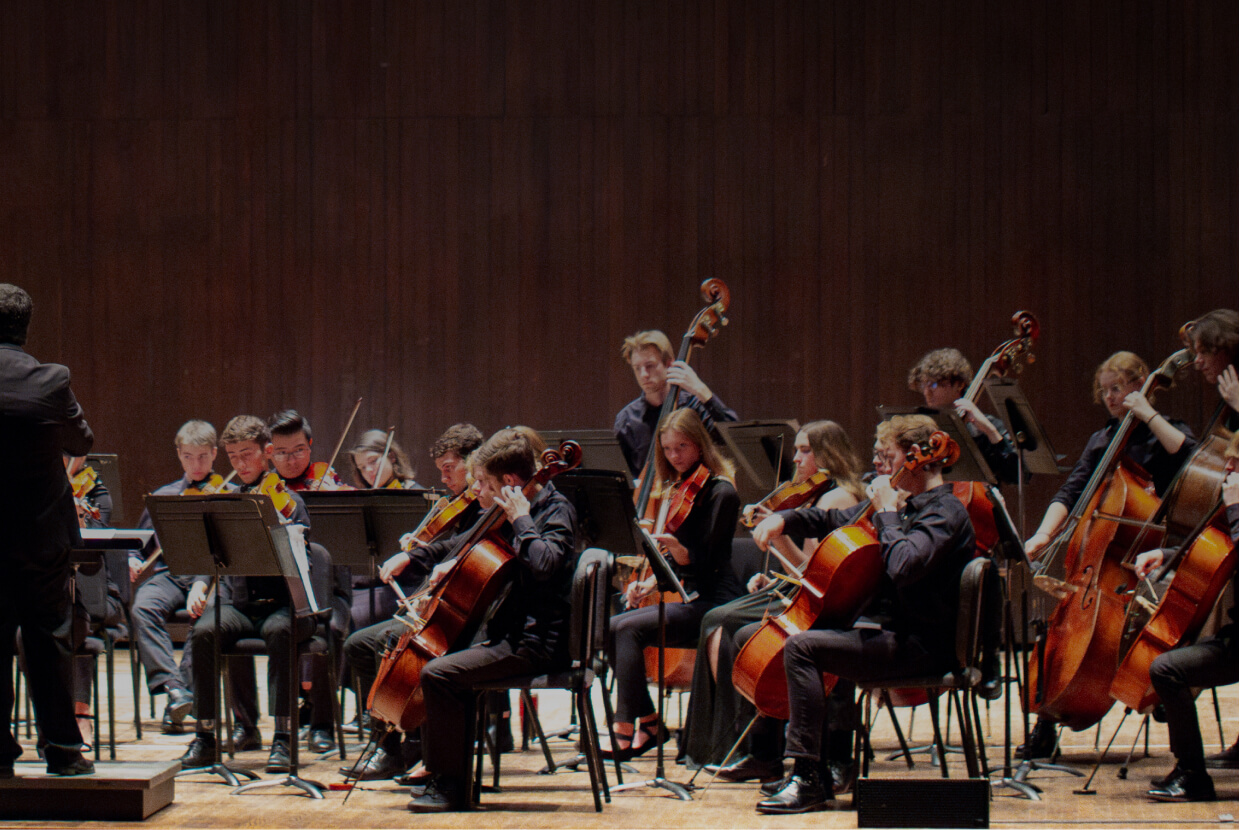 A group of musicians performs on a dimly lit stage. The ensemble includes various string instruments such as cellos and double basses. The musicians are seated and focused on their instruments, while the conductor directs them with raised hands.