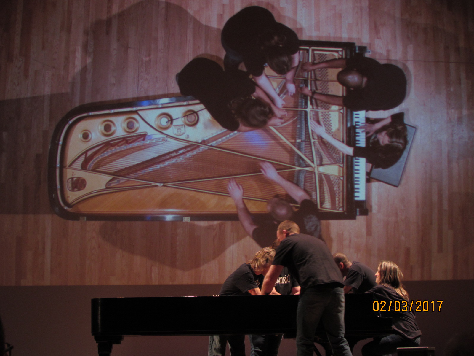 A group of people, viewed from above, huddles around the strings of a grand piano, hands positioned inside, possibly playing or tuning it. A large projection of the scene is displayed on the wall behind them. The date "02/03/2017" is shown in the lower right corner.