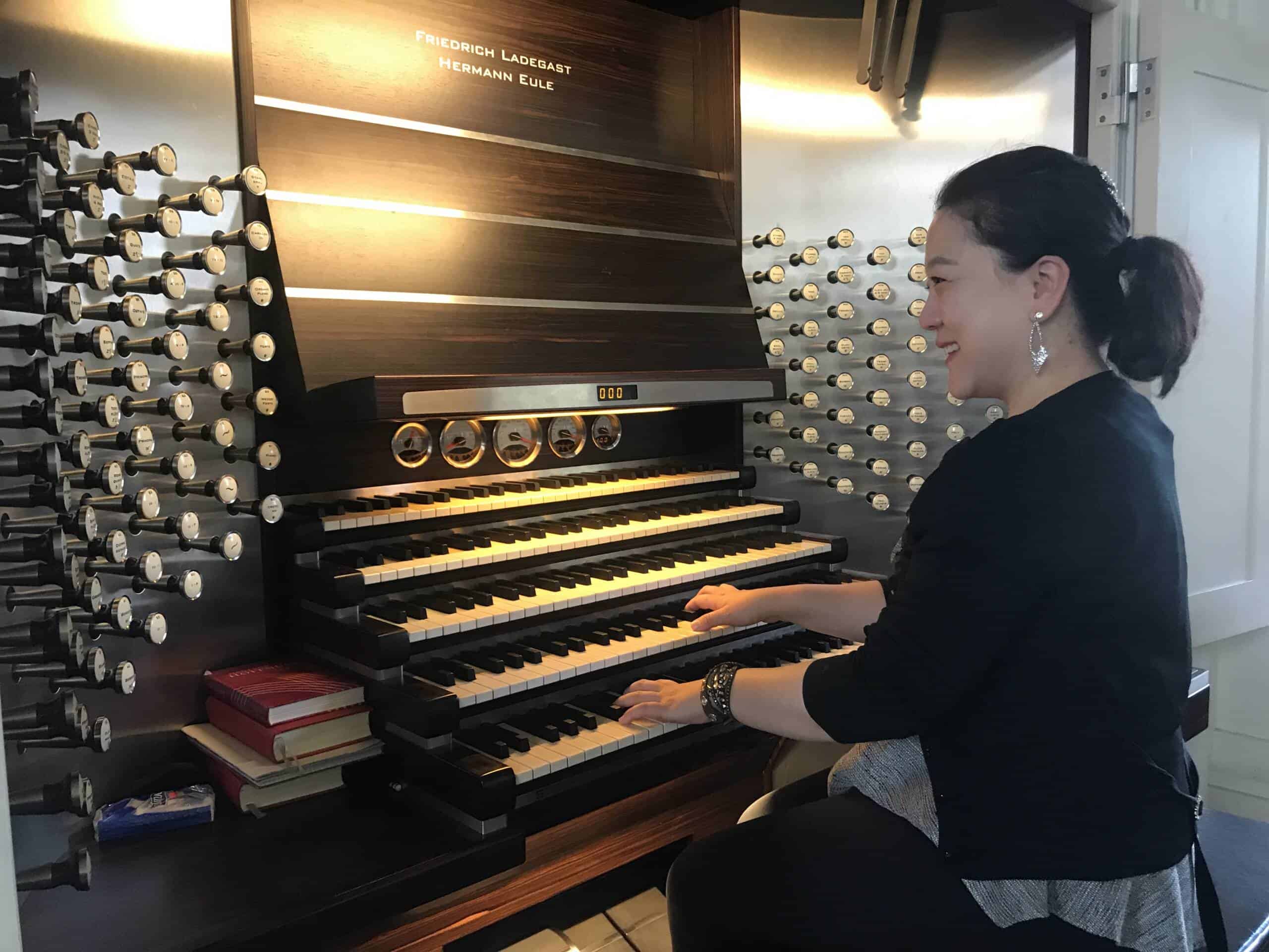A woman smiles while playing a large pipe organ with multiple keyboards, surrounded by rows of stops and sheet music on the side. The organ bears the name "Friedrich Ladegast Hermann Eule.