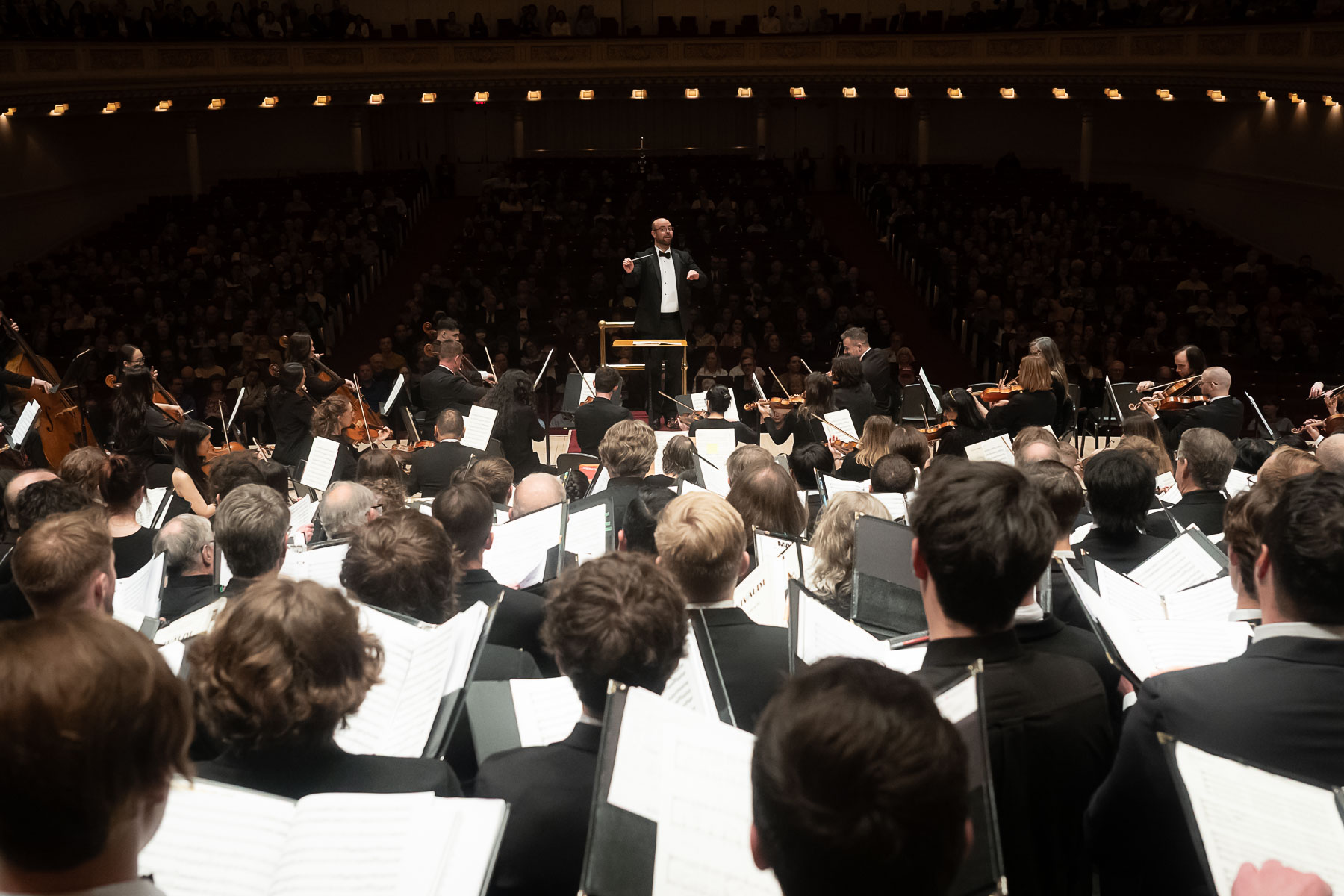 A large orchestra and choir perform on stage in a concert hall, with a conductor leading them. The audience is seated in the background, and the musicians and singers hold sheet music.