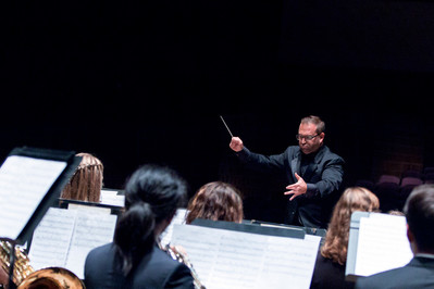 A conductor leads an orchestra during a performance, gesturing with a baton. Musicians with sheet music are visible in the foreground, and the stage is dimly lit.