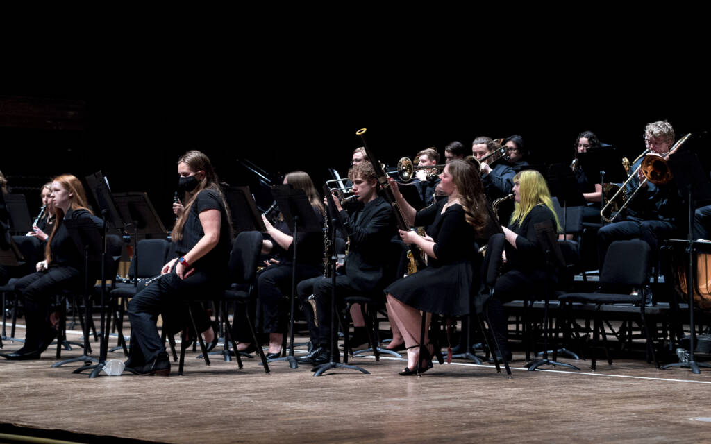 A group of musicians in black attire sit on stage with wind instruments, preparing to perform. Some are adjusting their music stands, and one person wears a black face mask. The stage is dimly lit.