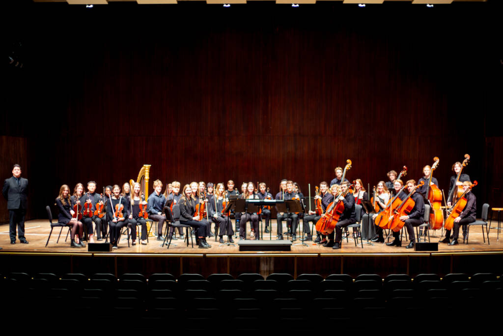A full orchestra of musicians with various string and brass instruments sits and stands on a stage in front of an empty audience, with a conductor standing to the left.