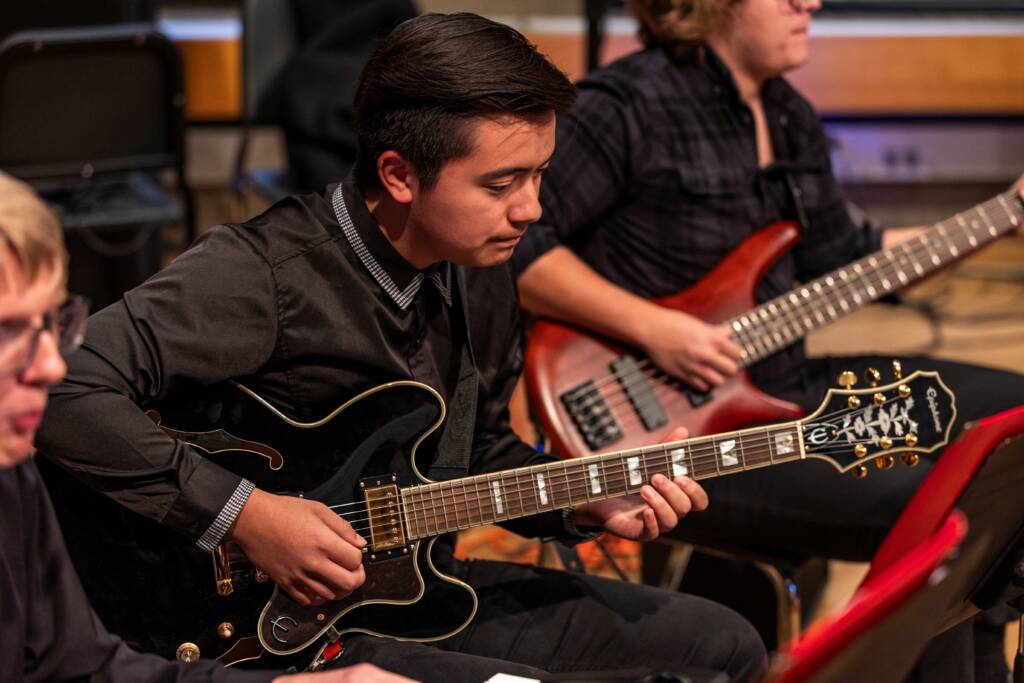 A young musician in a black shirt plays an electric guitar during a performance, with another person playing a bass guitar in the background.