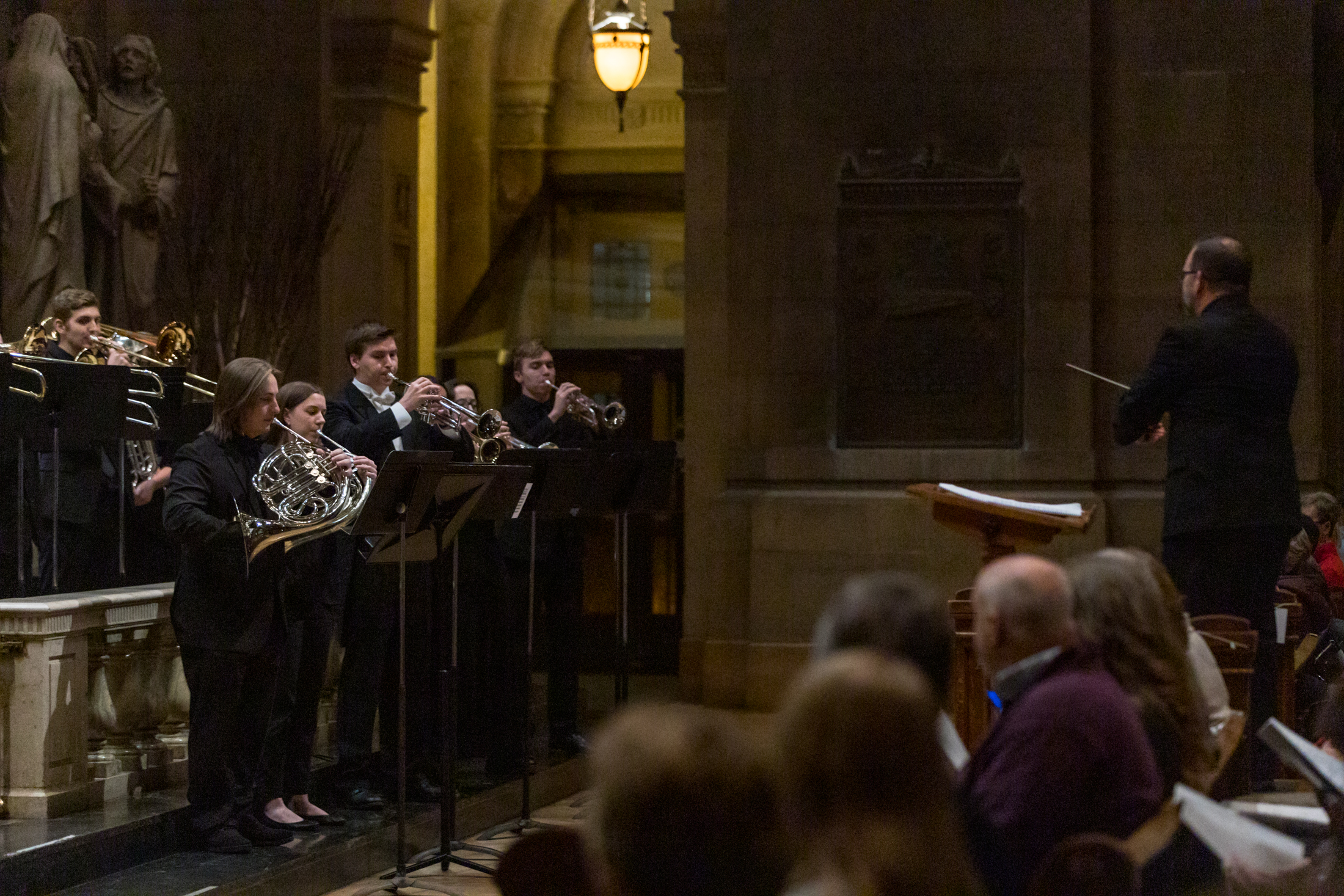A group of musicians plays French horns in a dimly lit church, facing a conductor who stands at a podium. Audience members are seated in the foreground, and statues and arches are visible in the background.