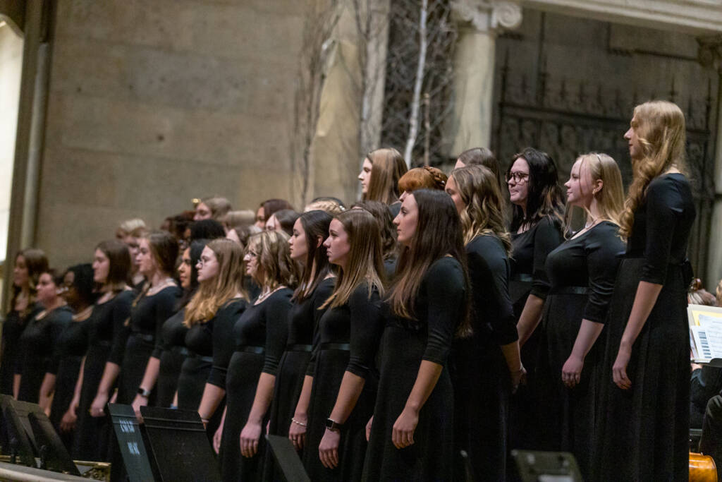 A large group of women in black dresses stands in rows, singing in a choir performance inside a grand hall with stone walls and columns.