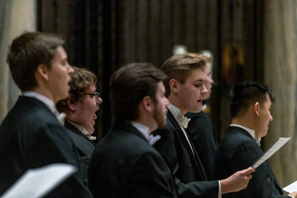 A group of young men dressed in tuxedos sing in a choir, holding sheet music. The focus is on the singers in profile, performing indoors with a blurred background.