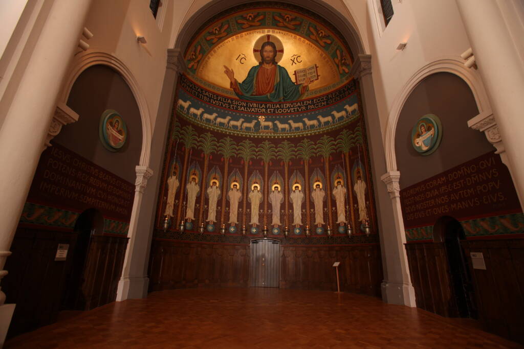 An ornate church apse features a large golden mosaic of Jesus with outstretched arms above a row of saintly figures, flanked by arched walls with religious text and circular icons, and a wooden floor below.